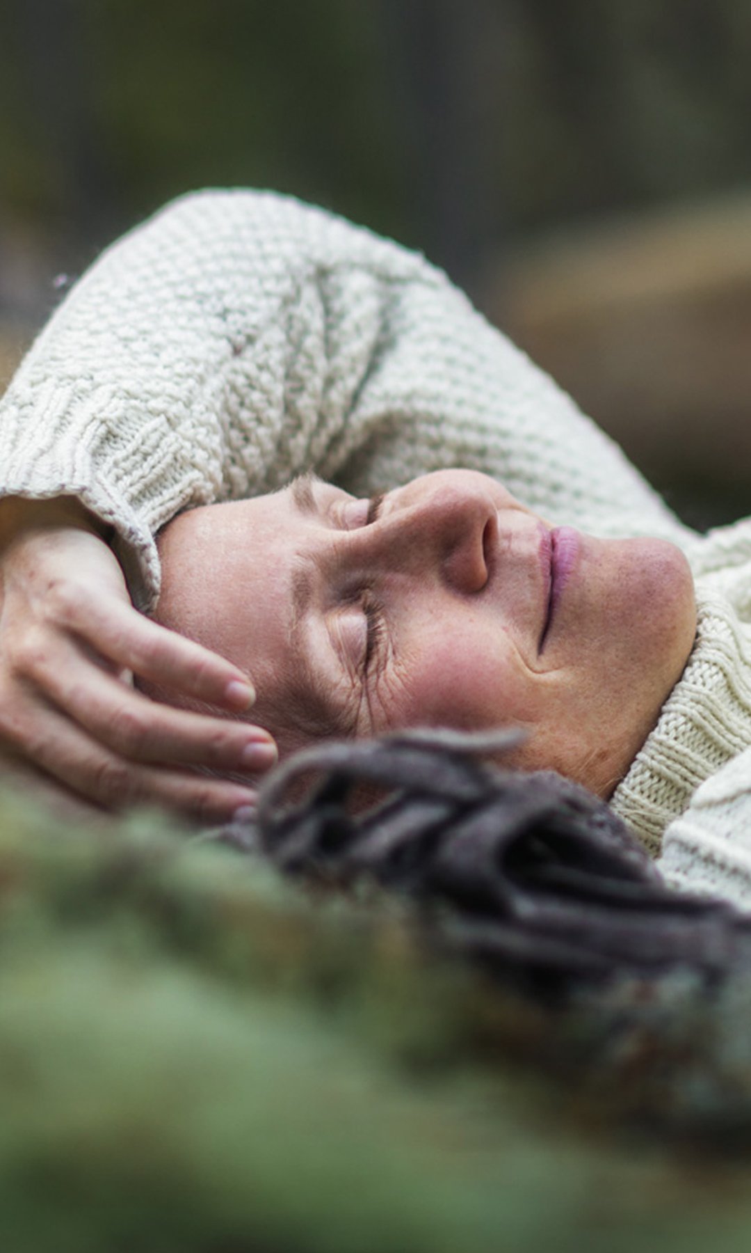 Kvinne med brystkreft med spredning ligger i mose på bakken i skogen. Hun har lukket øynene og hviler seg mens hun smiler. Illustrasjonsfoto
