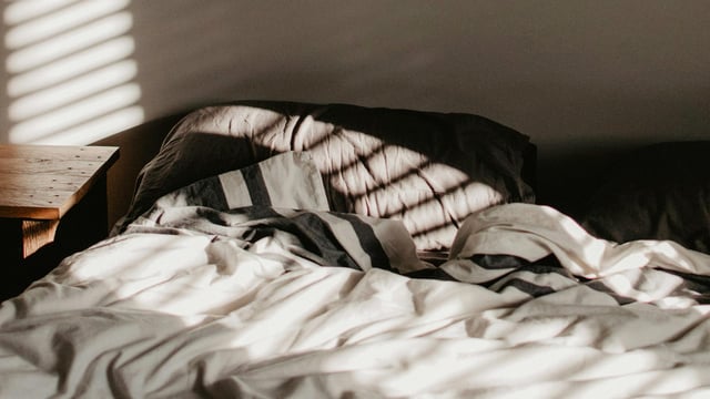 Uoppredd seng med lys fra åpne persienner som lyser opp sengetøy med duse farger. Illustrasjonsfoto.