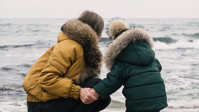 Brystkreftrammet kvinne holder hånden til et barn. De har varme tykke jakker på og sitter på huk ved vannkanten og ser ut over havet. Illustrasjonsfoto