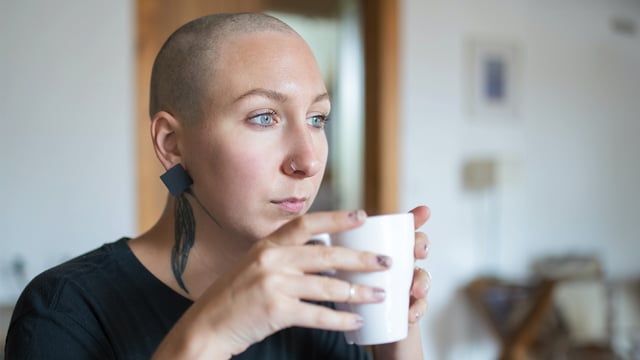 Nærbilde av en ung brystkreftrammet kvinne som holder en kopp. Kvinnen har barbert vekk håret. Hun ser tomt ut i rommet. Illustrasjonsfoto