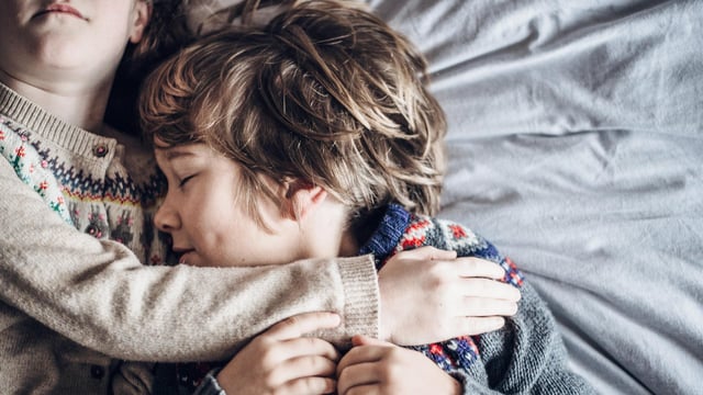 Nærbilde av et søskenpar som holder om hverandre i en seng. Begge har de strikkegensere på og gutten lukker øynene og ligger tett inntil sin søster. Illustrasjonsfoto