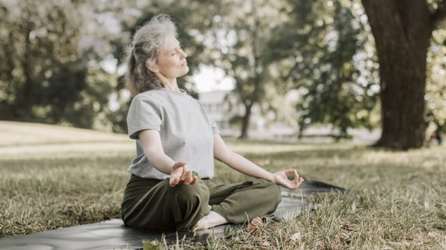 Kvinne med brystkreft med spredning som sitter i en park og praktiserer yoga. Hun har lukkede øyne. Illustrasjonsfoto