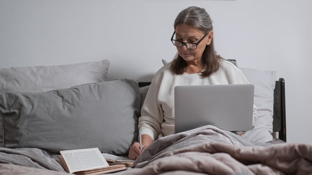 Brystkreftrammet kvinne sitter i sengen og jobber på en bærbar PC. Hun har bøker liggende ved siden av seg og tar notater. Illustrasjonsfoto