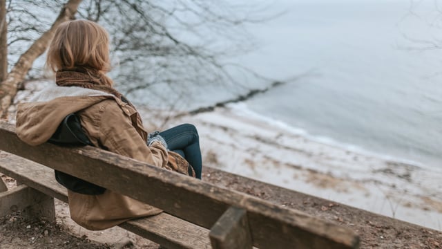 Kvinne som er rammet av tretthet fra kreftsykdom som sitter på en benk og ser ut over vannet på høsten. Illustrasjonsfoto
