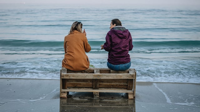 To kvinner sitter på en benk ved vannkanten på en strand og snakker sammen. Det er høst i luften og de har varme jakker på. Illustrasjonsfoto