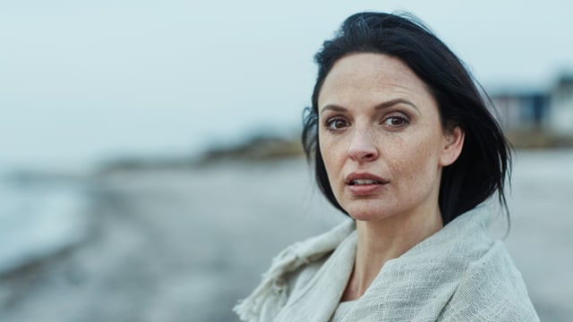 Mørkhåret kvinne med brystkreft med spredning som står på stranden og ser inn i kameraet. Illustrasjonsfoto