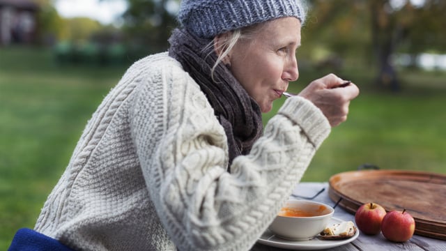 Kvinne med MBC sitter med lue og strikkegenser ved et bord utendørs og spiser suppe i høstvær. Illustrasjonsfoto.