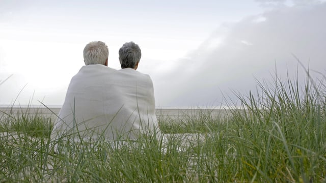 Et eldre par, hvor kvinnen er rammet av brystkreft, sitter sammen under et teppe på stranden og ser ut over havet. Vi ser dem bakfra. Illustrasjonsfoto