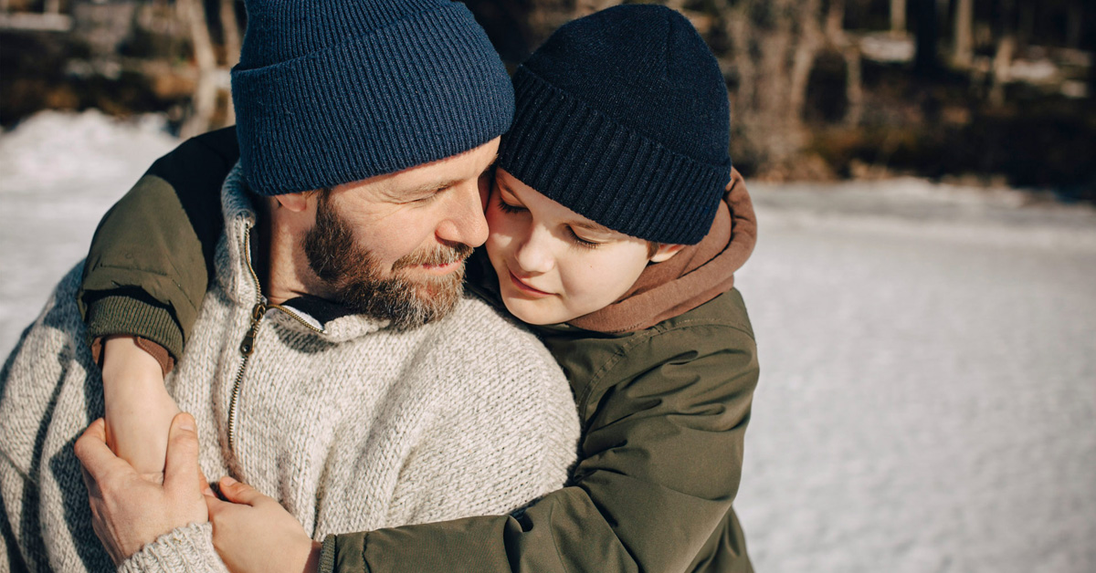 En ung gutt søker trøst og støtte hos faren sin, de er utendørs om vinteren. Foto.