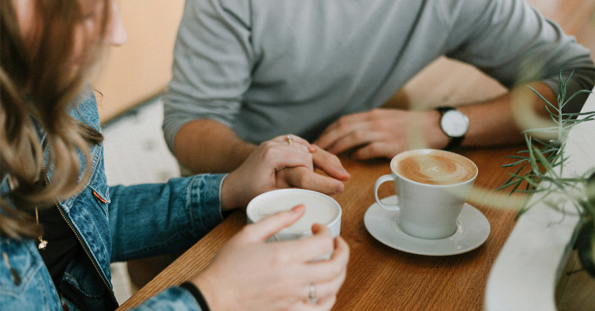 Nærbilde av en likeperson som holder en støttende hånd over samtalepartnerens hånd, de sitter på en kafé med hver sin kaffe
